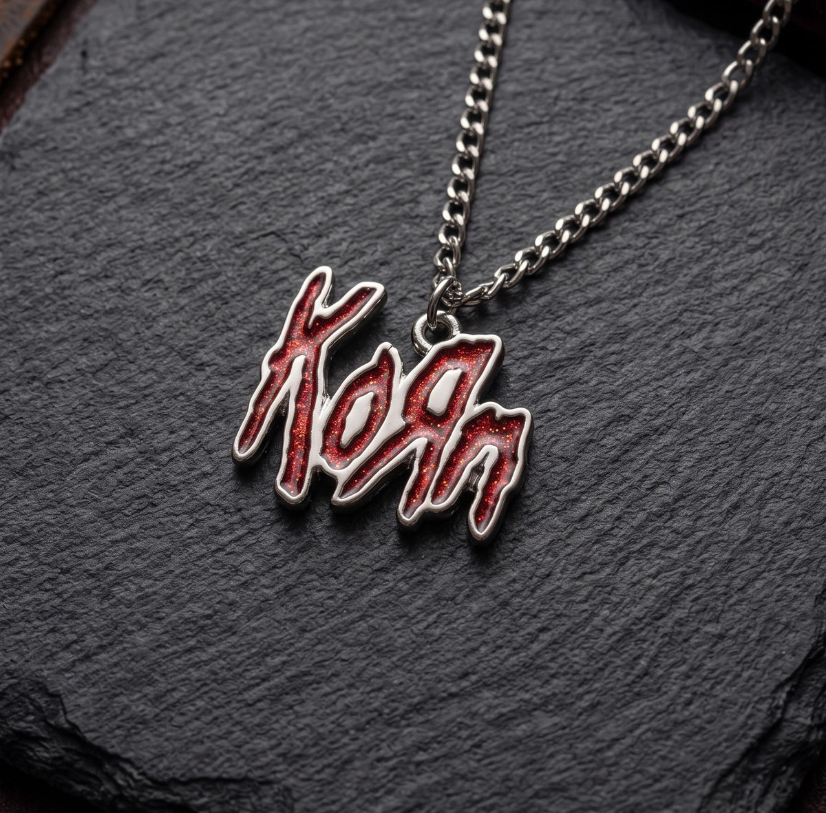 We Rock Korn red glitter logo pendant on a silver-tone metal chain resting on a textured black slate surface